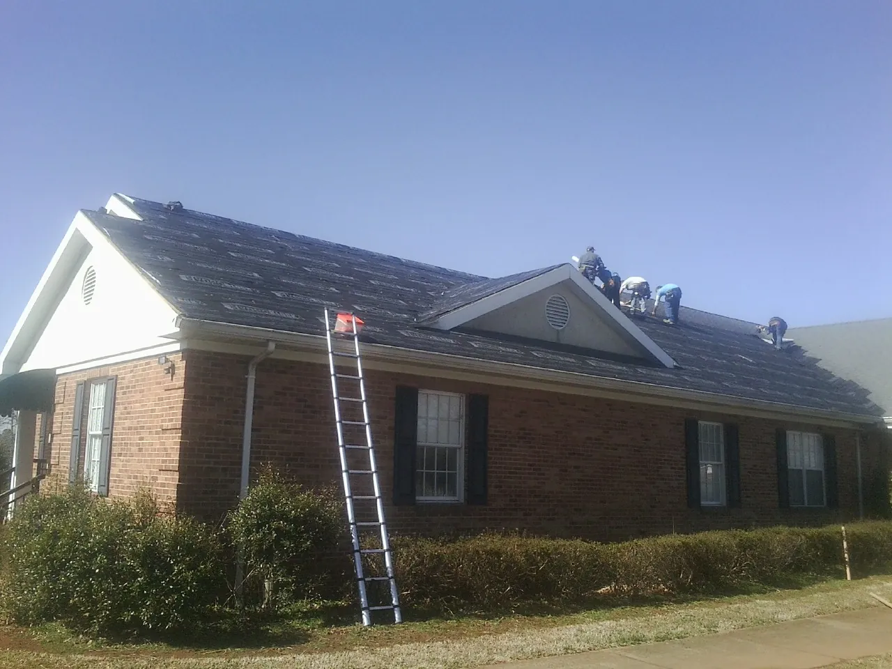 Crew tearing shingles off a brick ranch roof with a ladder set — Metal Roof Repair work by our crew in Chittenango